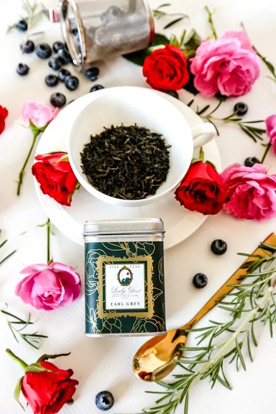 Earl Grey Premium Flavored Black Tea in a white cup with a green tea caddy in front, surrounded by red and pink roses, blueberries, and a golden spoon