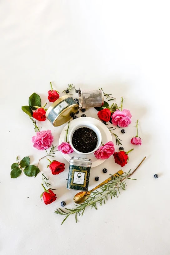 English Breakfast Premium Loose Leaf Tea in a white cup surrounded by gold and green tea caddies, pink and red roses, and a golden spoon on a white background