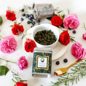 Premium Sencha Green Loose Leaf Tea in a green tin caddy and white cup sorrounded with red and pink roses, golden spoon, and blueberries