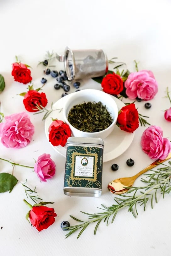 Premium Sencha Green Loose Leaf Tea in a green tin caddy and white cup sorrounded with red and pink roses, golden spoon, and blueberries